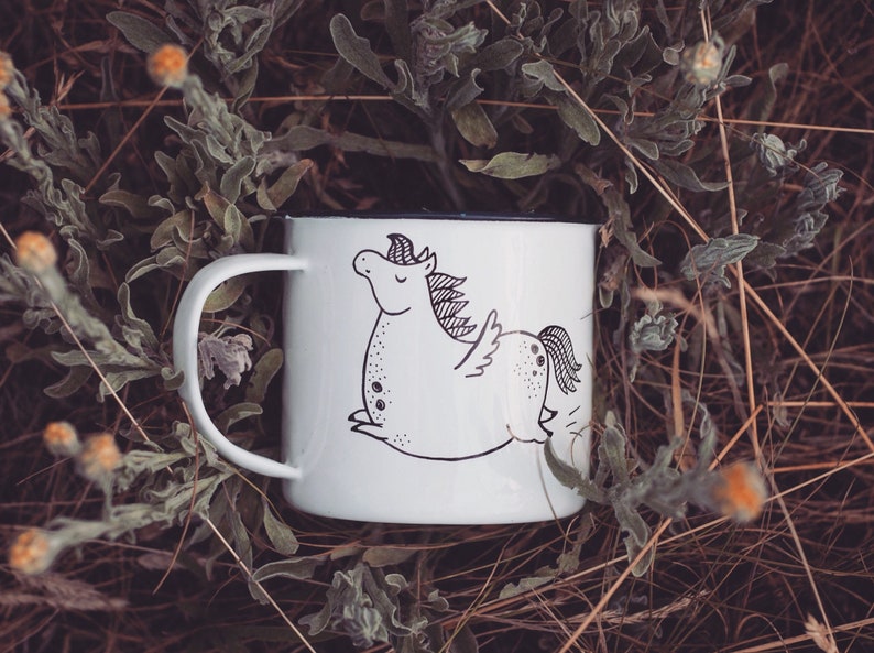 May include: White enamel mug with a black and white illustration of a winged unicorn. The mug is sitting on a bed of dried grasses and flowers.