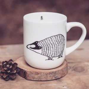 May include: White ceramic mug with a black and white illustration of a badger. The mug has a small tree design on the inside.