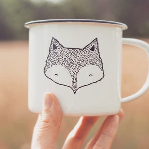 Peut inclure: Une tasse en émail blanche avec une illustration en noir et blanc d'un visage de renard.