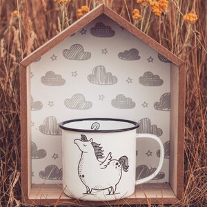 May include: White enamel mug with a black rim and a black and white illustration of a unicorn with a cloud background. The mug is sitting in a wooden house-shaped frame.