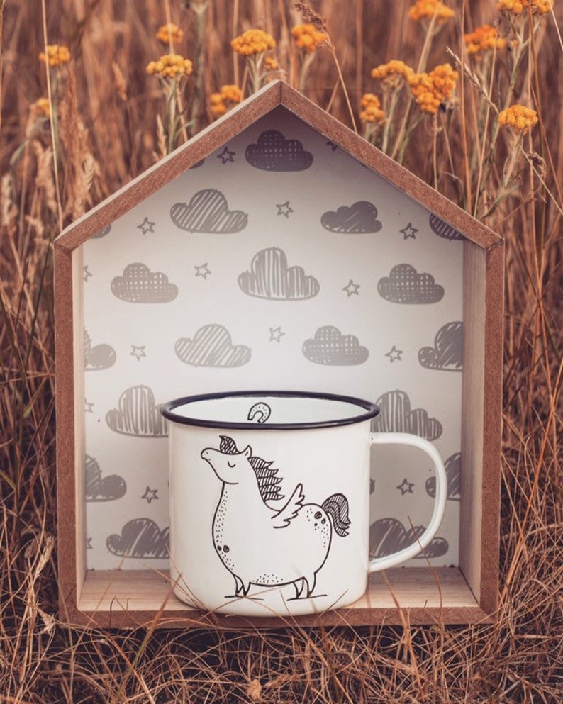 May include: White enamel mug with a black rim and a black and white illustration of a unicorn with a cloud background. The mug is sitting in a wooden house-shaped frame.