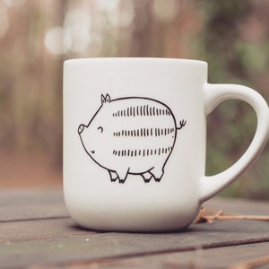 Könnte beinhalten: Weiße Keramiktasse mit einer schwarz-weißen Illustration eines Schweins auf der Vorderseite.