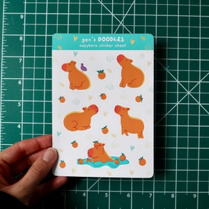 Peut inclure: Une feuille d'autocollants avec des illustrations de capybaras dans différentes poses, notamment en train de nager, de s'asseoir et de se tenir debout. La feuille est blanche avec une bordure bleue et le texte "gen's DOODLES capybara sticker sheet" en haut.