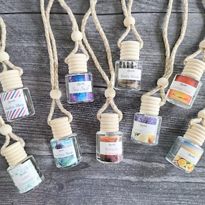 May include: Nine different car air fresheners hanging from strings. The air fresheners are in clear glass bottles with colorful labels and wooden stoppers. The labels have different scents, including Star Stuff, Rustic Wood, Summer Days, Barber Shop, Electric Blue, Lavender Citrus, Mango Tea, Odor Eliminator, and Citrus.