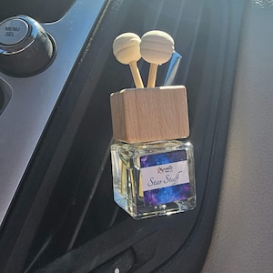 May include: A car air freshener with a wooden top and two spherical wooden diffusers. The glass container has a label that reads "Moonfit Star Stuff" with a galaxy design. The freshener is placed in a car's air vent.