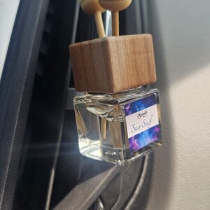 Puede incluir: Un ambientador para coche con una parte superior de madera y dos difusores de bola de madera. El frasco de vidrio transparente tiene una etiqueta con un dise&ntilde;o de galaxia y el texto "Star Stuff". El ambientador est&aacute; sujeto a una rejilla de ventilaci&oacute;n del coche.