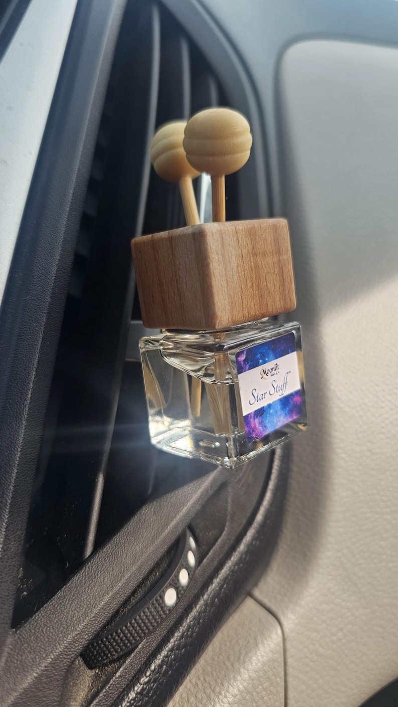 Puede incluir: Un ambientador para coche con una parte superior de madera y dos difusores de bola de madera. El frasco de vidrio transparente tiene una etiqueta con un dise&ntilde;o de galaxia y el texto "Star Stuff". El ambientador est&aacute; sujeto a una rejilla de ventilaci&oacute;n del coche.