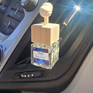 Puede incluir: Un ambientador para coche con tapa de madera y botella de cristal. La botella tiene una etiqueta que dice "Moonlit Star Stuff" con un dise&ntilde;o de galaxia. El ambientador est&aacute; colocado en una rejilla de ventilaci&oacute;n del coche.