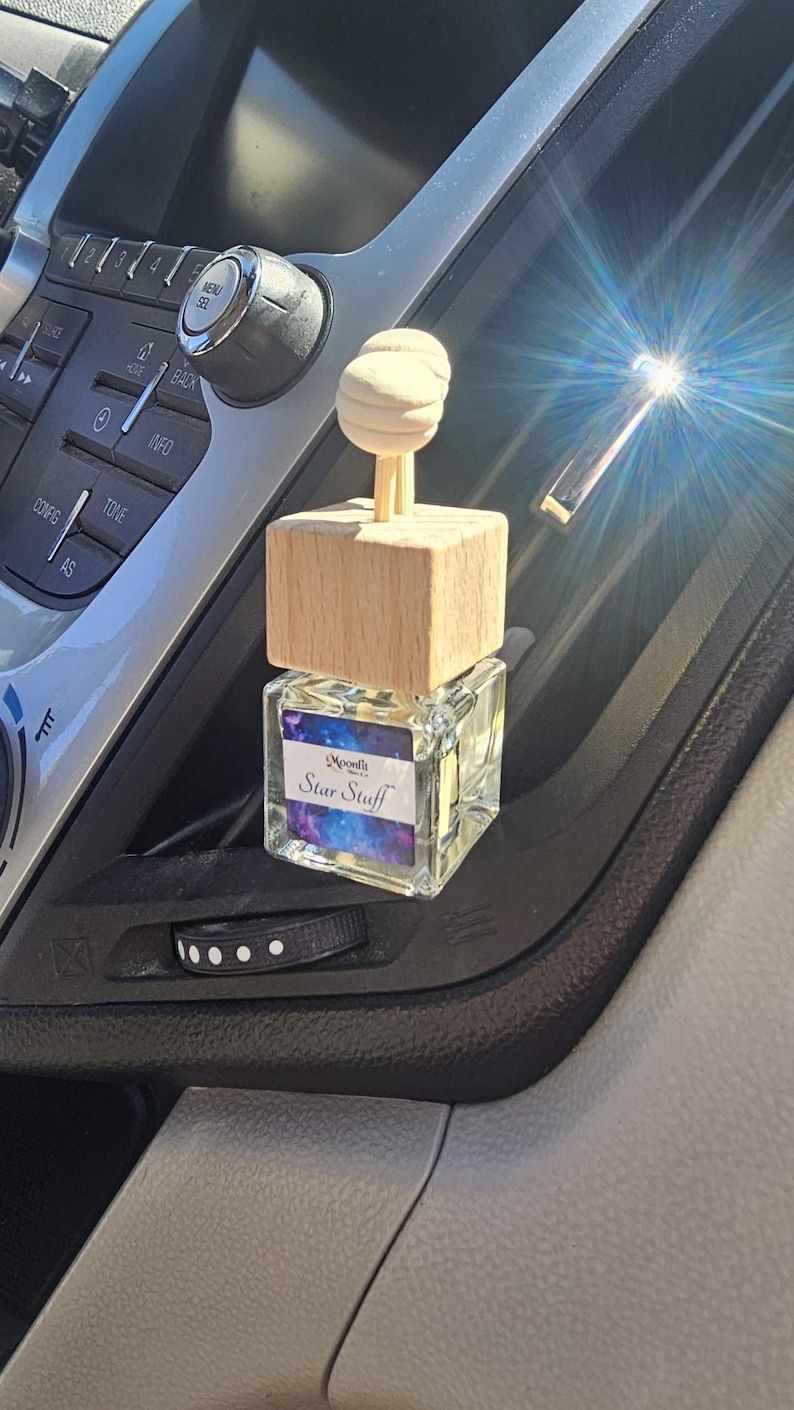 Puede incluir: Un ambientador para coche con tapa de madera y botella de cristal. La botella tiene una etiqueta que dice "Moonlit Star Stuff" con un dise&ntilde;o de galaxia. El ambientador est&aacute; colocado en una rejilla de ventilaci&oacute;n del coche.