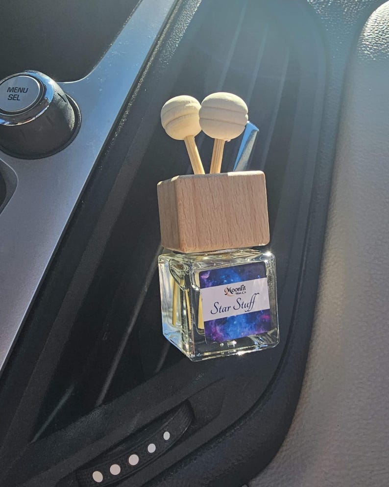 Puede incluir: Un ambientador para coche con una parte superior de madera y dos difusores esf&eacute;ricos de madera. El recipiente de vidrio tiene una etiqueta que dice "Moonfit Star Stuff" con un dise&ntilde;o de galaxia. El ambientador est&aacute; colocado en una rejilla de ventilaci&oacute;n del coche.