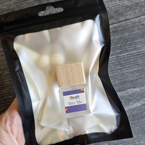 Puede incluir: Una peque&ntilde;a botella de vidrio transparente con tapa de madera, etiquetada "Bite Me" en texto blanco sobre fondo morado. La botella est&aacute; dentro de una bolsa de pl&aacute;stico negro con interior plateado. La imagen est&aacute; tomada sobre una superficie de madera.