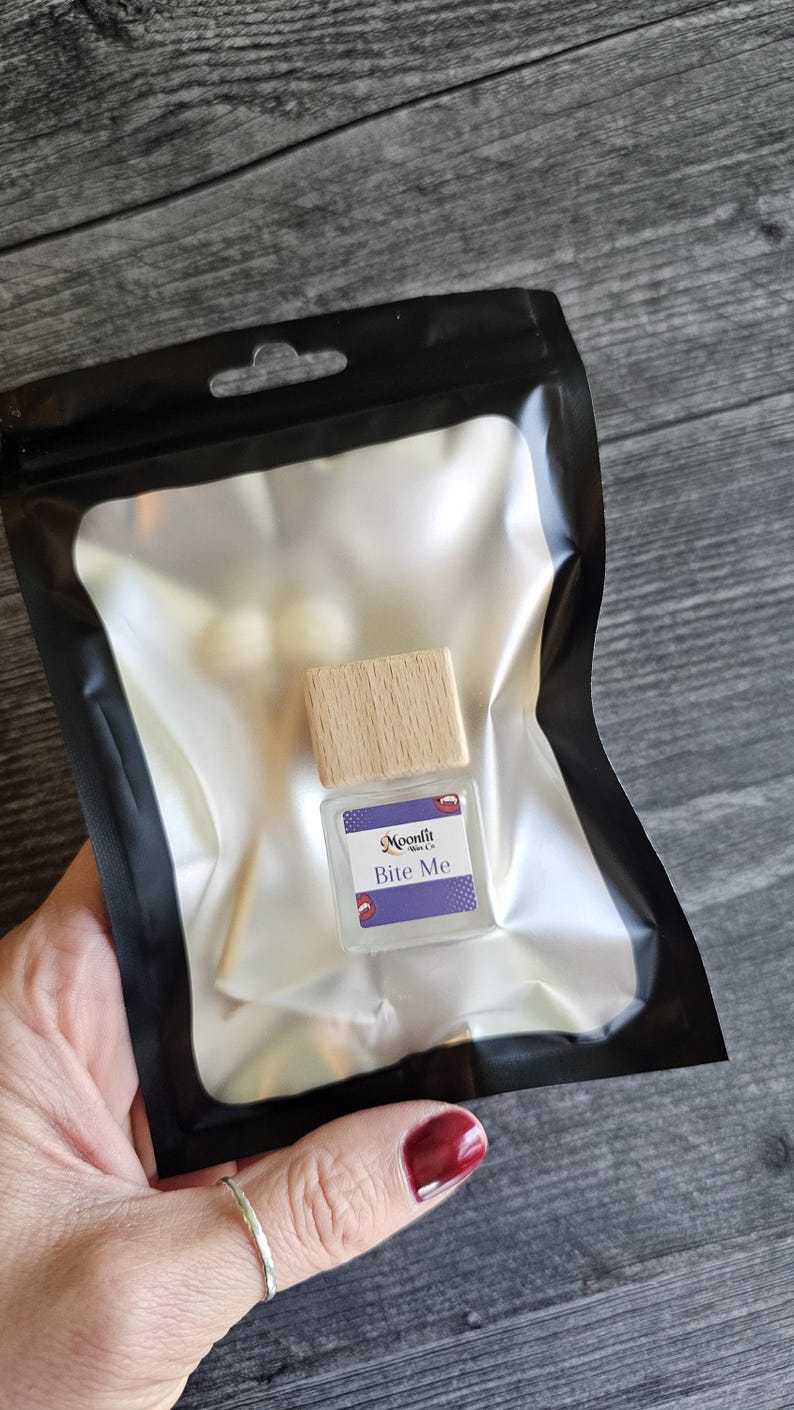 Puede incluir: Una peque&ntilde;a botella de vidrio transparente con tapa de madera, etiquetada "Bite Me" en texto blanco sobre fondo morado. La botella est&aacute; dentro de una bolsa de pl&aacute;stico negro con interior plateado. La imagen est&aacute; tomada sobre una superficie de madera.