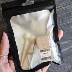 Puede incluir: Una botella de vidrio transparente con tapa de madera y dos palitos de madera, empaquetada en una bolsa de pl&aacute;stico negra. La botella tiene una etiqueta que dice "Moonfit Apricot Grove". La bolsa se sostiene en una mano.