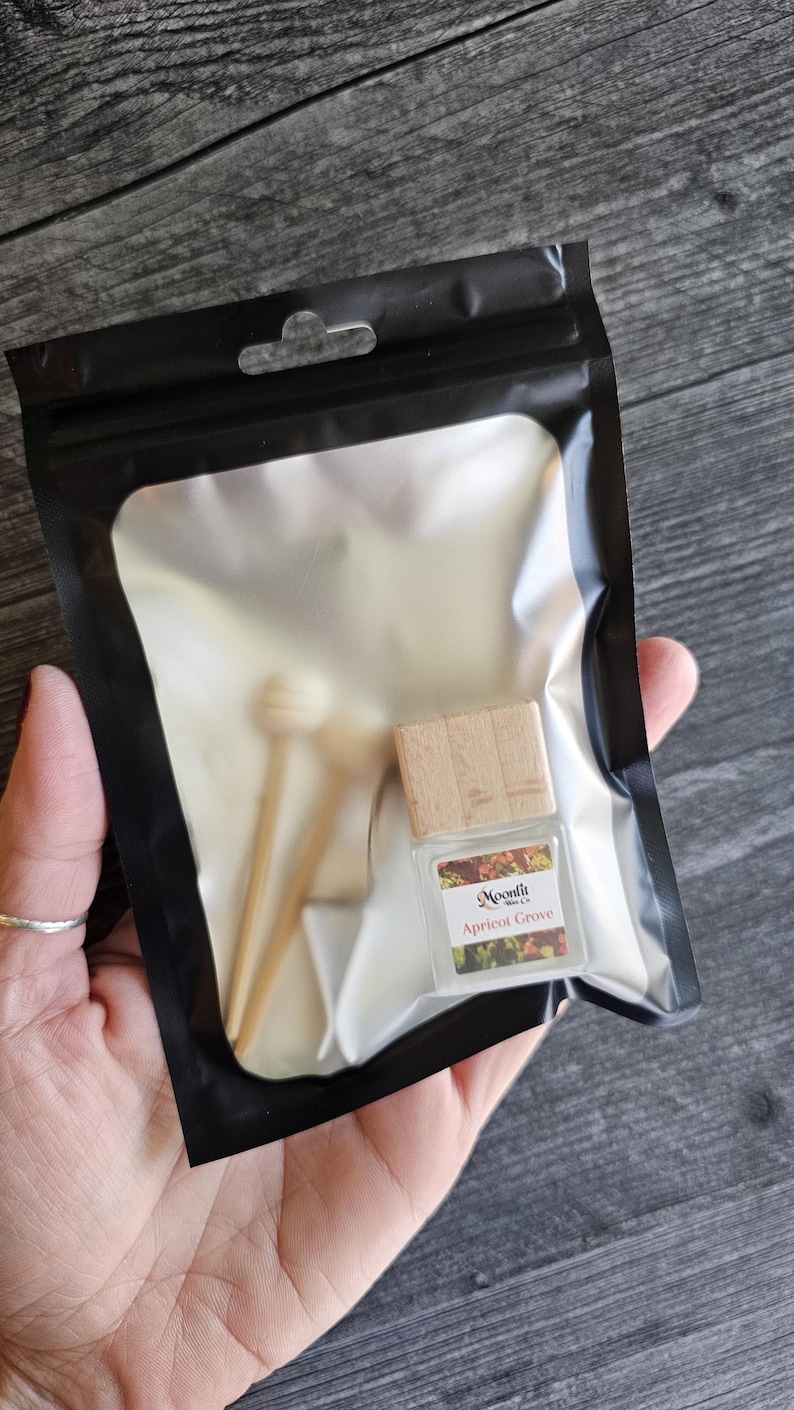 Puede incluir: Una botella de vidrio transparente con tapa de madera y dos palitos de madera, empaquetada en una bolsa de pl&aacute;stico negra. La botella tiene una etiqueta que dice "Moonfit Apricot Grove". La bolsa se sostiene en una mano.