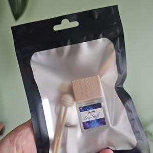 Puede incluir: Una botella transparente y cuadrada con una tapa de madera y una etiqueta que dice "Star Stuff" est&aacute; dentro de una bolsa de pl&aacute;stico transparente con un borde negro. La bolsa tambi&eacute;n contiene dos palitos de madera y una peque&ntilde;a cuchara de madera. El fondo es verde claro.