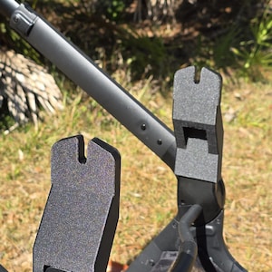 Könnte beinhalten: Nahaufnahme eines schwarzen Kinderwagenrahmens mit strukturierter Oberfläche. Der Rahmen hat ein einzigartiges Design mit einem gebogenen Griff und einer Stützstruktur. Der Hintergrund zeigt grünes Gras und Blätter.