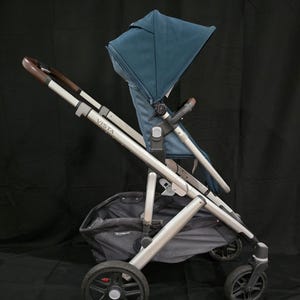 Puede incluir: Un cochecito plateado y gris con una capota azul. El cochecito tiene una cesta grande debajo y la palabra "VISTA" está impresa en el manillar.