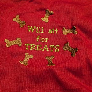 May include: Red fabric with embroidered text and dog bone designs. The text reads "Will sit for TREATS" in green thread, surrounded by brown dog bone shapes. The fabric has a textured appearance.