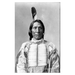 May include: Black and white portrait of a Native American man wearing traditional clothing and a feather headdress. He has a serious expression on his face.