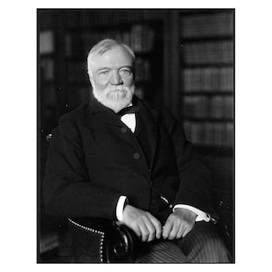 May include: A black and white portrait of a man with a white beard and mustache, wearing a dark suit and a white shirt with a bow tie. He is sitting in a chair with a bookshelf in the background.