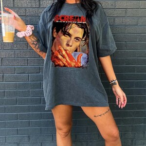 May include: Dark gray t-shirt featuring a graphic from the movie "Scream." The graphic shows a close-up of a person with blood-covered hands. The word "SCREAM" is printed in red at the top, with the text "WE ALL DO A LITTLE MAD SOMETIMES" below.