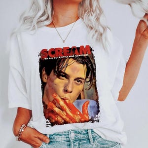 May include: White t-shirt featuring a graphic from the movie "Scream." The graphic includes the title in red letters and an image of a person with blood on their hands. The text "WE ALL GO A LITTLE MAD SOMETIMES" is also included.