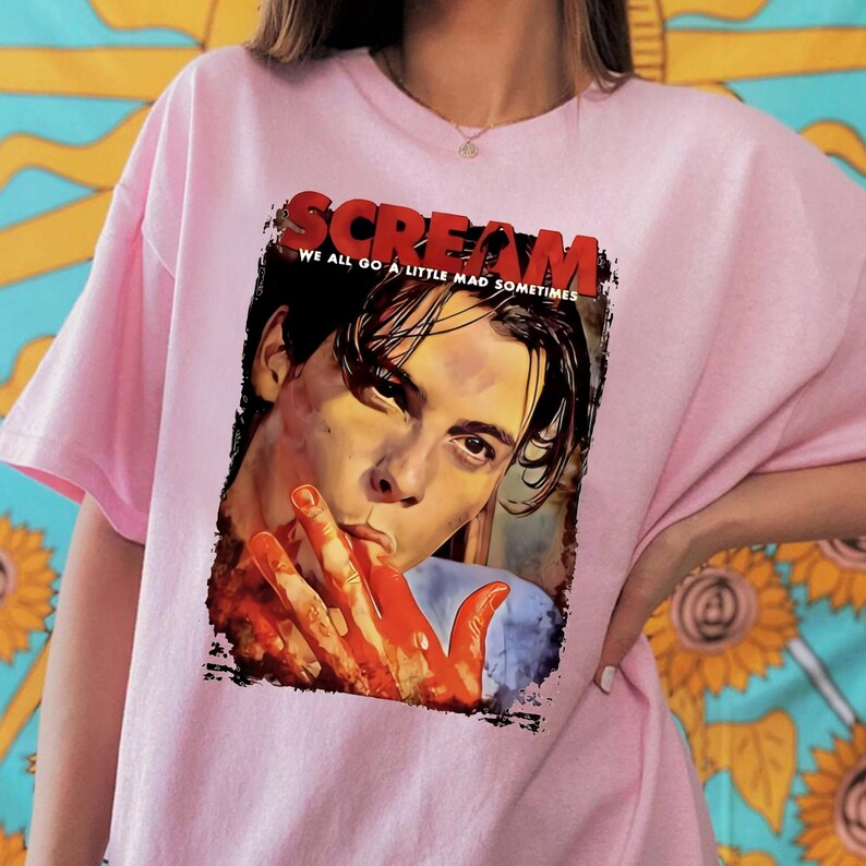 May include: Pink t-shirt featuring a graphic from the movie "Scream." The graphic includes the word "Scream" in red, the phrase "We all go a little mad sometimes," and a portrait of a person with blood on their hands.