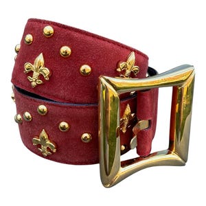 May include: A red suede belt featuring gold-toned metal fleur-de-lis emblems and round studs. The belt is secured with a large, polished gold-toned square buckle that has a smooth, reflective finish and a prominent prong mechanism.