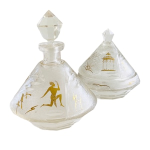 May include: A set of two clear glass perfume bottles with gold accents. The bottles are shaped like pyramids and have a geometric design. The bottle on the left has a figure of a person in a kneeling position, while the bottle on the right has a pagoda design.