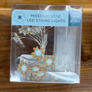 May include: Clear plastic package containing Missouri Star LED string lights. The lights are shaped like stars with orange, teal, and yellow accents. The package indicates the lights are 7 ft (2.13m) long.