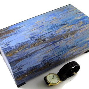 Puede incluir: Una caja de madera azul y dorada con vetas de m&aacute;rmol con un reloj de cuero negro en la parte superior.
