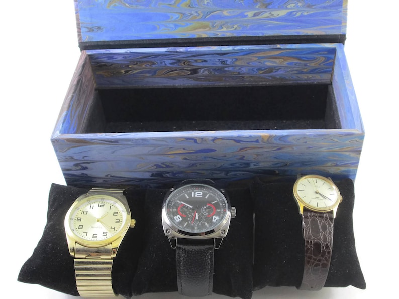 Puede incluir: Una caja de madera de color azul y dorado con vetas de m&aacute;rmol y un interior de terciopelo negro. La caja contiene tres relojes de pulsera, uno con una correa dorada y una esfera blanca, uno con una correa negra y una esfera blanca y uno con una correa de cuero marr&oacute;n y una esfera blanca.
