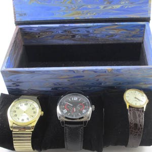 Puede incluir: Una caja de madera de color azul y dorado con vetas de m&aacute;rmol y un interior de terciopelo negro. La caja contiene tres relojes de pulsera, uno con una correa dorada y una esfera blanca, uno con una correa negra y una esfera blanca y uno con una correa de cuero marr&oacute;n y una esfera blanca.
