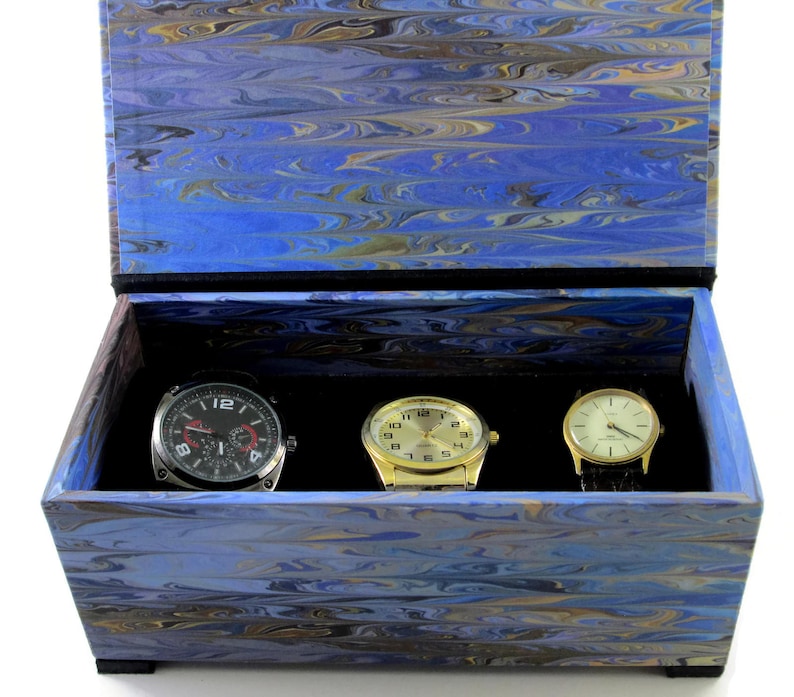 Puede incluir: Una caja de madera azul y dorada con vetas de m&aacute;rmol y un interior de terciopelo negro. La caja contiene tres relojes de pulsera: uno plateado con esfera negra, uno dorado con esfera blanca y uno dorado con esfera negra.