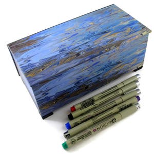 Puede incluir: Una caja de madera azul y dorada con vetas de m&aacute;rmol, con una tapa con bisagras, que contiene un juego de seis marcadores negros de punta fina. Los marcadores est&aacute;n etiquetados con "MICRON 03" y "TOUCH OF COLOR" en letras negras.