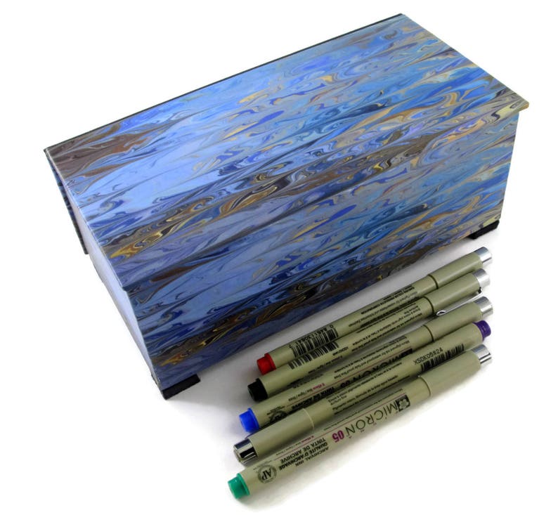 Puede incluir: Una caja de madera azul y dorada con vetas de m&aacute;rmol, con una tapa con bisagras, que contiene un juego de seis marcadores negros de punta fina. Los marcadores est&aacute;n etiquetados con "MICRON 03" y "TOUCH OF COLOR" en letras negras.