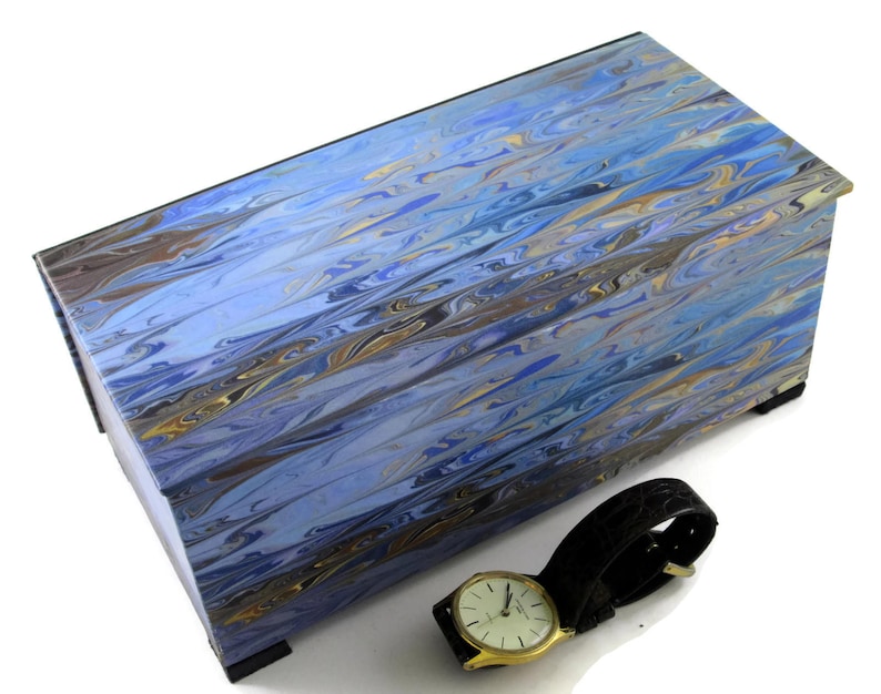 Puede incluir: Una caja de madera azul y dorada con vetas de m&aacute;rmol con un reloj de cuero negro en la parte superior.