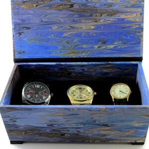 Puede incluir: Una caja de madera azul y dorada con vetas de m&aacute;rmol y un interior de terciopelo negro. La caja contiene tres relojes de pulsera: uno plateado con esfera negra, uno dorado con esfera blanca y uno dorado con esfera negra.