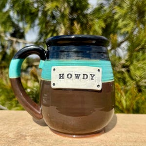 Peut inclure: Mug en céramique faite à la main avec une base marron, une bande turquoise et un bord noir. La tasse présente une anse avec des accents turquoise et noirs. Le mot "HOWDY" est affiché sur une plaque rectangulaire blanche.