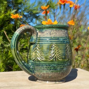 Puede incluir: Una taza de cerámica con un esmalte verde brillante que presenta una textura moteada y un patrón tallado de árboles de hoja perenne alrededor del centro. La taza tiene un asa grande y curva y una base ancha, con una flor naranja en el borde.
