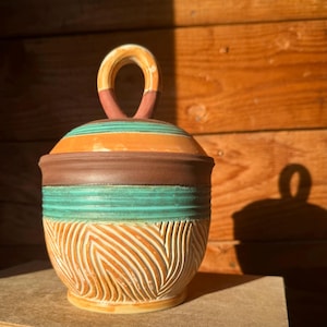 Może przedstawiać: Ręcznie robiony ceramiczny słoik z pokrywką. Słoik ma rzeźbiony, teksturowany wzór w odcieniach pomarańczu i bieli. Pokrywka jest w paski z pomarańczowymi, turkusowymi i brązowymi paskami. Uchwyt to pętla z pomarańczowymi i różowymi akcentami. Słoik to dekoracyjny pojemnik do przechowywania.