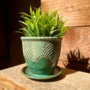 May include: A decorative ceramic planter with a green and blue glaze, filled with vibrant green artificial grass. The planter has a textured, carved design and sits on a matching saucer. A stylish accent for home decor.