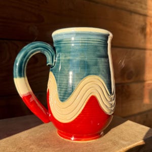 May include: Handmade ceramic mug with a large handle. The mug features a vibrant design with a red base, a wavy cream-coloured band, and a blue top. The handle transitions from red to cream to blue.