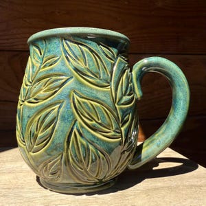 May include: A ceramic mug with a glossy finish in shades of teal, forest green, and lime green. The exterior features a relief pattern of overlapping leaves carved into the surface. It has a wide, rounded body and a curved handle on the right side.