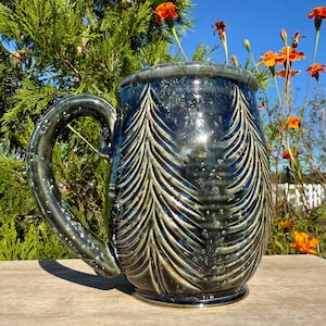 May include: A ceramic mug with a glossy dark blue and green glaze featuring a carved pattern of vertical pine trees. The mug has a large, curved handle and a smooth, reflective surface with subtle light-colored speckles throughout the finish.