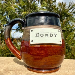 May include: A handcrafted ceramic mug with a black rim and handle, featuring a brown and tan body. The mug has a white rectangular label with the word "HOWDY" in black letters. The mug is designed for hot beverages.