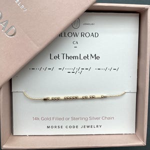 May include: A gold-colored Morse code necklace with the message "Let Them Let Me" on a white card inside a pink box. The necklace is made of small beads and is labeled as 14k gold filled or sterling silver.