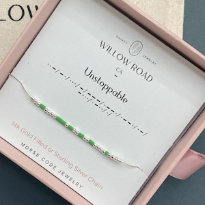 May include: A delicate silver chain necklace with green and silver beads spelling "Unstoppable" in Morse code. The necklace is presented in a pink box with the Willow Road Jewelry logo and text.