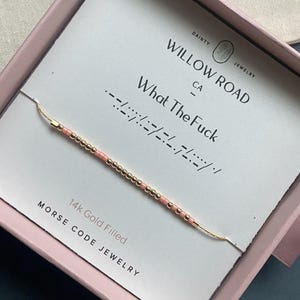 Peut inclure: Un bracelet en code Morse rempli d'or avec des perles roses, présenté dans une boîte rose. La carte indique "What The Fuck" en code Morse, avec le nom de la marque "Willow Road Jewelry" et le texte "14k Gold Filled".