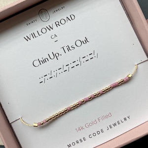 May include: A gold-filled Morse code bracelet with pink and gold beads, presented in a pink box. The card reads "Willow Road CA" and "Chin Up, Tits Out". The bracelet is labeled as 14k Gold Filled Morse Code Jewelry.